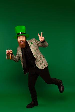 Excited bearded man in hat holding bottles of beer and showing peace sign on green backgroundの写真素材