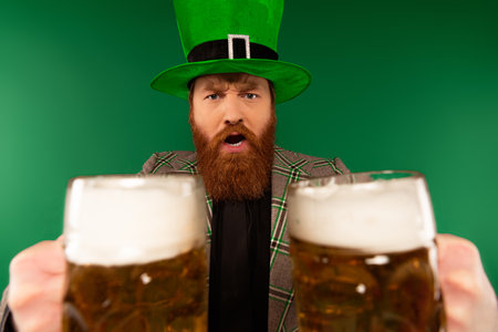 Serious bearded man in hat holding glasses of beer isolated on greenの写真素材