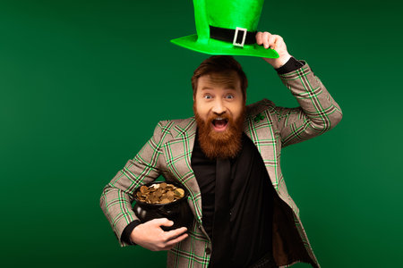 Excited bearded holding hat and pot with coins during saint patrick day isolated on greenの写真素材