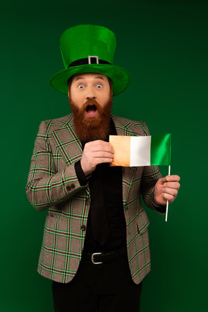 Shocked bearded man in hat holding Irish flag isolated on greenの写真素材