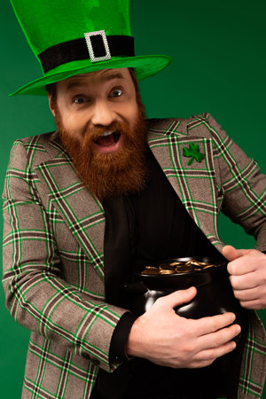 Amazed man in hat with clover holding pot with golden coins isolated on greenの写真素材