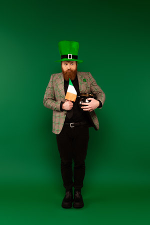 Pensive man in hat holding Irish flag and pot with coins on green backgroundの写真素材