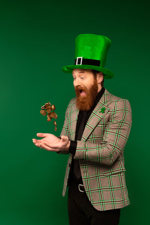 Shocked bearded man in hat throwing coins during saint patrick day isolated on greenの写真素材