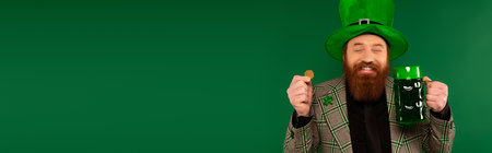 Joyful man in hat with clover holding coin and glass of beer isolated on green, banner