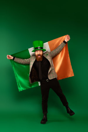 Full length of excited bearded man in hat holding Irish flag on green backgroundの写真素材