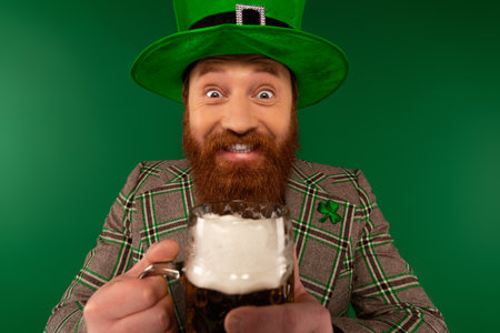 Cheerful bearded man in hat holding beer and looking at camera during saint patrick day isolated on greenの写真素材