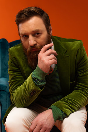 Trendy bearded model in velour jacket sitting on armchair on red background