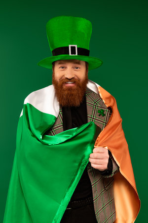 Smiling man in hat with clover wrapping in Irish flag isolated on greenの写真素材