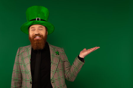 Cheerful man in hat with clover pointing with hand isolated on greenの写真素材