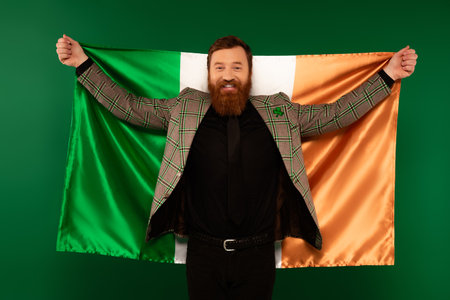 Smiling bearded man with clover on jacket holding Irish flag isolated on greenの写真素材