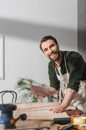 Smiling carpenter in apron holding sandpaper near board and looking at camera in workshopの写真素材