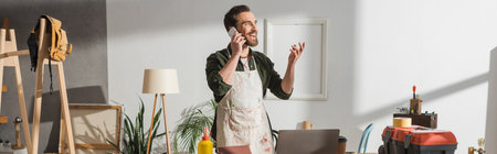 Smiling craftsman talking on cellphone near tools and laptop in studio, banner