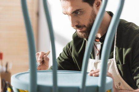 Bearded craftsman in apron painting on wooden chair in workshop