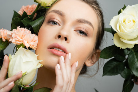 portrait of sensual woman touching face while looking at camera near roses and carnations isolated on greyの写真素材