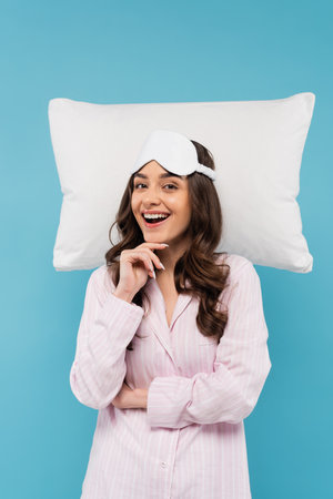 excited young woman in pajamas and night mask smiling near white flying pillow isolated on blueの写真素材