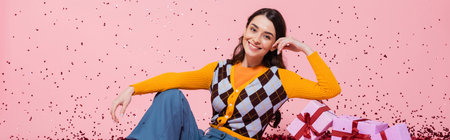 cheerful woman in fashionable attire posing near gift boxes and festive confetti on pink background,...