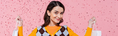 brunette woman in stylish attire holding shopping bags and smiling at camera on pink background,...