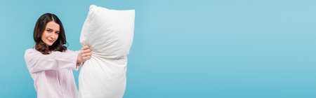 brunette young woman in pajamas holding white pillow isolated on blue, bannerの写真素材