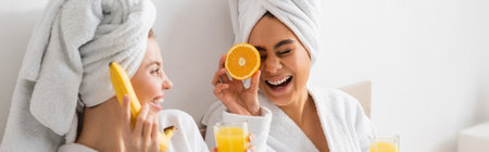cheerful african american woman covering eye with ripe orange while having fun near friend with banana and juice, bannerの写真素材