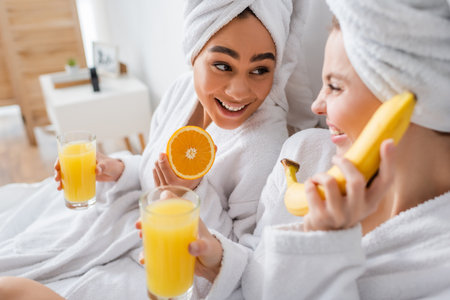 happy african american woman holding ripe orange and juice near blurred friend having fun in bedroomの写真素材