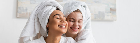 pretty and cheerful interracial women in white robes and towels smiling with closed eyes in bedroom, bannerの写真素材