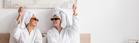 happy interracial women in white towels and stylish sunglasses toasting with champagne and looking at each other, bannerの写真素材