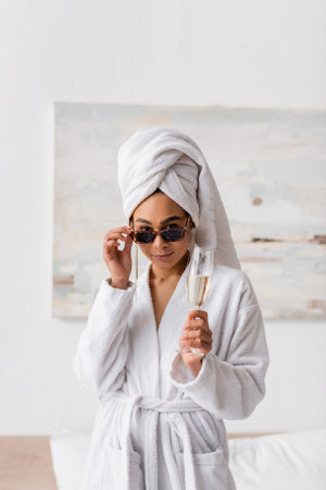 young african american woman in white bathrobe and towel holding champagne and looking at camera over sunglasses in bedroomの写真素材