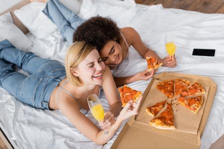high angle view of excited multiethnic women holding delicious pizza and cocktails while lying on bed at homeの写真素材