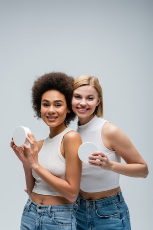 pretty multiethnic models in white tank tops posing with containers of cosmetic cream isolated on greyの写真素材