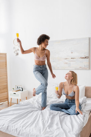 joyful african american woman in bra and jeans posing with cocktail near blonde friend sitting on bedの写真素材