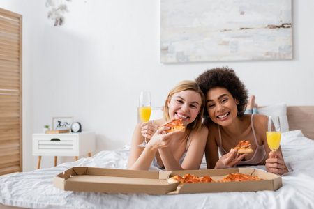 joyful multiethnic women looking at camera while lying on bed with delicious pizza and champagne glasses with cocktailsの写真素材