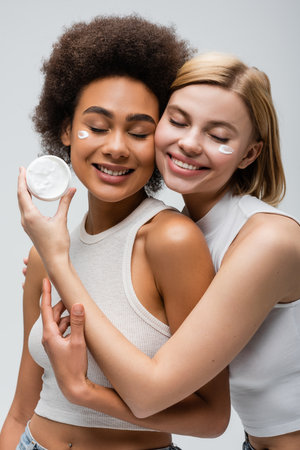 sensual blonde model holding face cream near smiling african american woman isolated on greyの写真素材