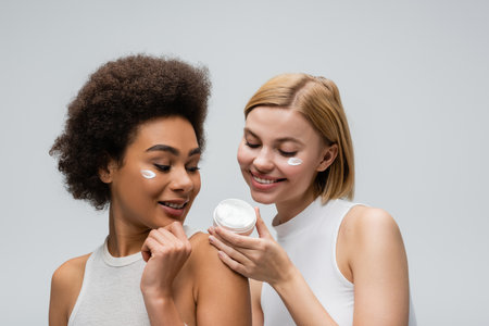 pleased multiethnic women in white tops looking at container with face cream isolated on greyの写真素材
