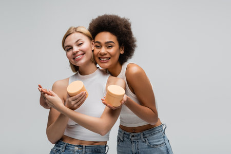 pleased multiethnic women holding hands while posing with cosmetic cream isolated on greyの写真素材