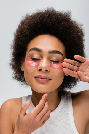 young and positive african american woman touching face with hydrogel eye patches isolated on greyの写真素材