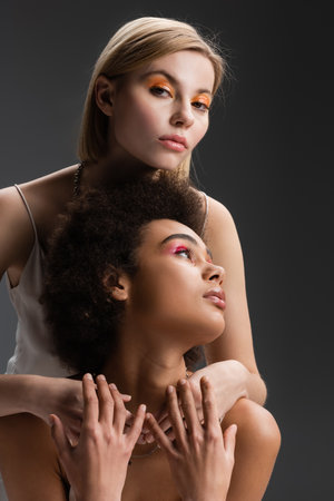 blonde model with orange eye shadows embracing brunette african american woman while looking at camera isolated on greyの写真素材