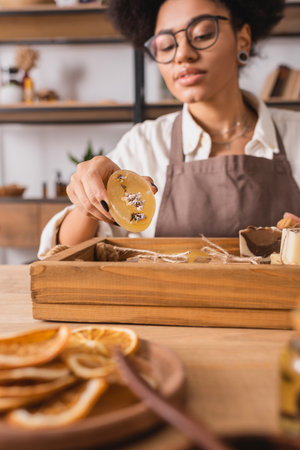 blurred african american woman in eyeglasses holding handmade soap near wooden tray in craft workshopの写真素材