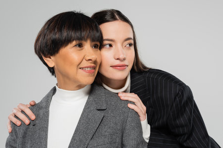 positive asian mother and daughter in white turtlenecks and blazers looking away isolated on greyの写真素材