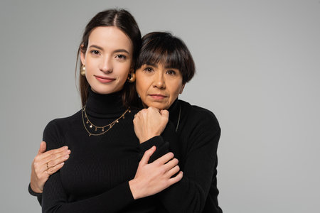 middle aged asian woman embracing young and trendy daughter while looking at camera isolated on grey