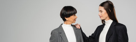 positive asian woman in stylish clothes touching shoulder of brunette mother isolated on grey,...