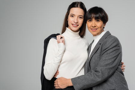 asian mother and daughter in elegant clothes embracing and looking at camera isolated on greyの写真素材