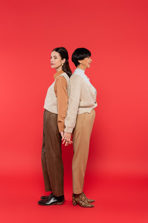 full length of asian mother and daughter in stylish casual attire holding hands while standing back...