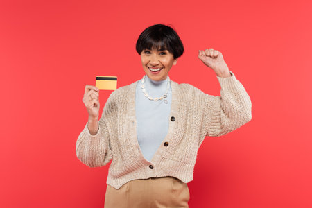 brunette asian woman in knitted cardigan holding credit card and celebrating success isolated on coralの写真素材