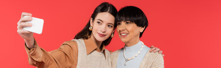 young asian woman in knitted vest taking selfie with happy mother isolated on coral, banner