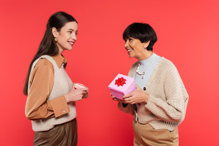 pleased asian mother and daughter in stylish clothes holding presents and looking at each other isolated on coralの写真素材