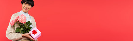 happy asian woman holding roses and presents isolated on coral, bannerの写真素材