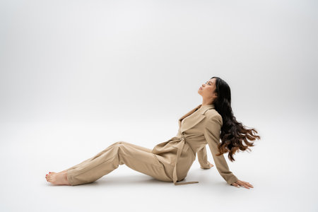 side view of barefoot asian model with long hair sitting in fashionable pantsuit on grey backgroundの写真素材