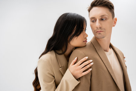 sensual asian woman with long brunette hair hugging man in beige jacket isolated on greyの写真素材