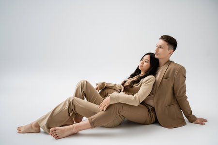 full length of barefoot interracial couple in beige pantsuits sitting on grey backgroundの写真素材