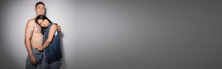 shirtless man with tattooed body embracing brunette asian woman in denim vest on grey background, bannerの写真素材